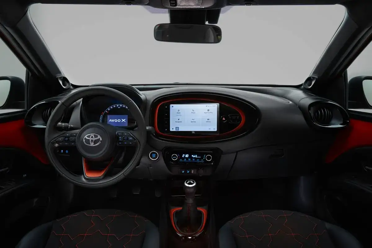 Interni moderni della Toyota Aygo X con volante multifunzione, schermo touchscreen centrale e dettagli rossi.