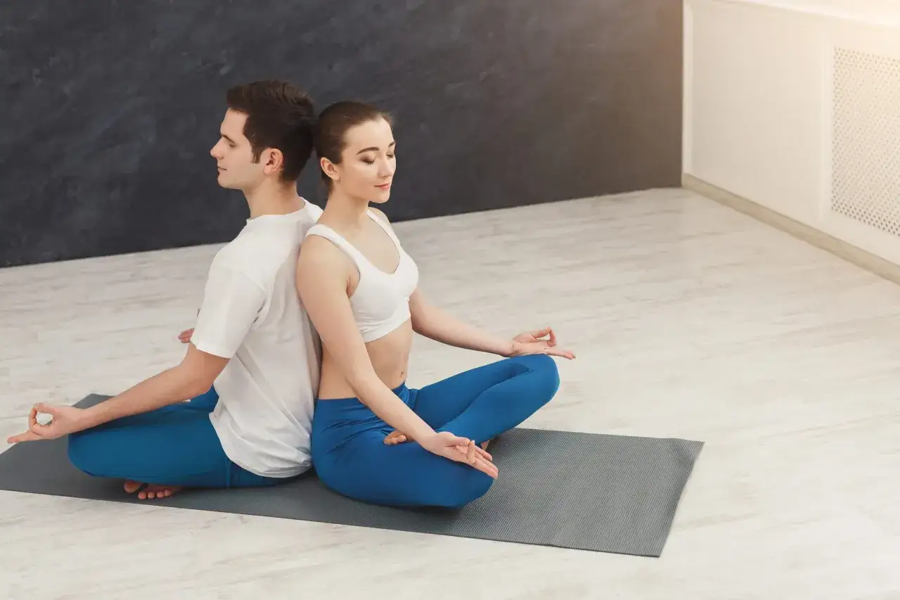 Coppia in meditazione, schiena contro schiena, in una posa che evoca il sesso tantrico, con le mani in mudra.