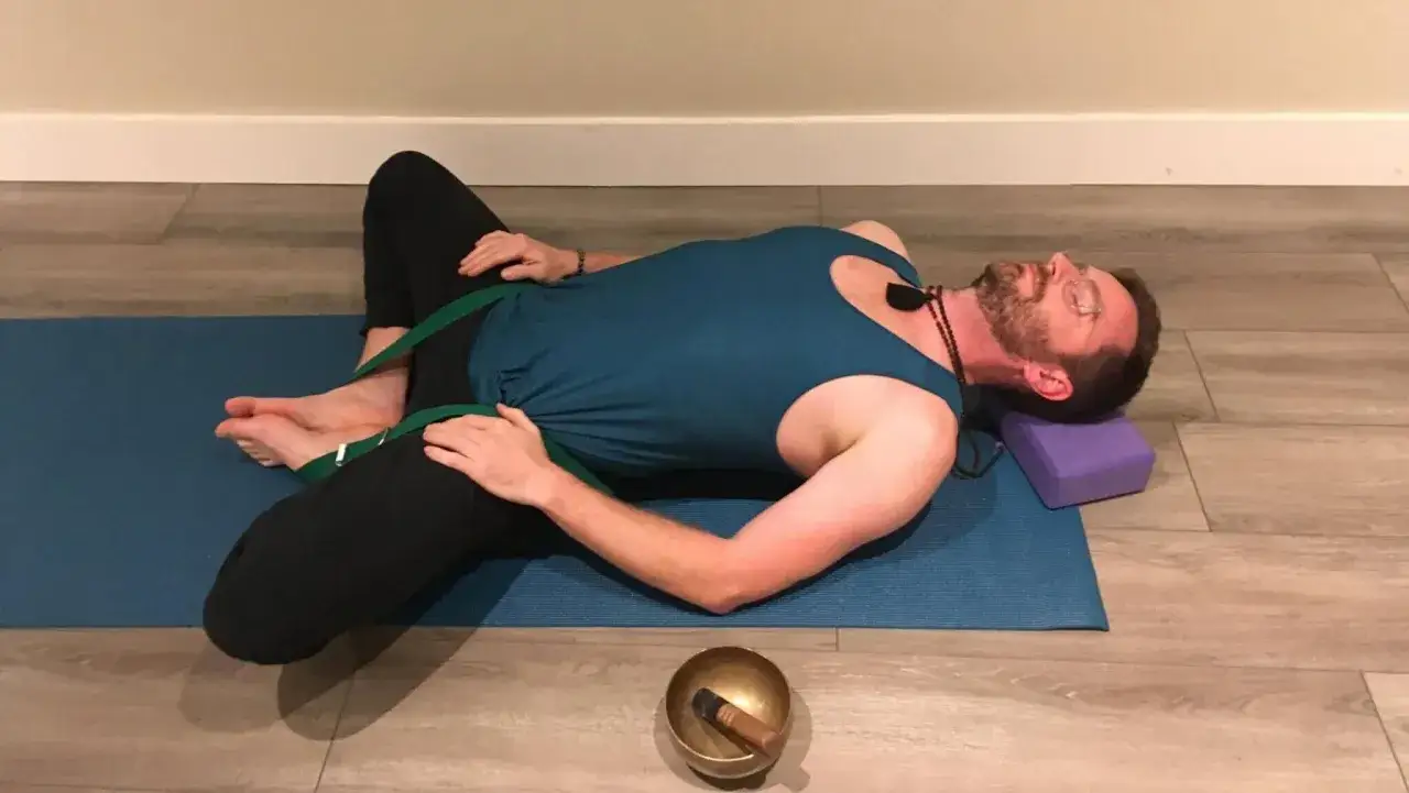 Uomo in posizione farfalla yoga, con cinghia per facilitare l'apertura delle anche. Un blocco viola sostiene la testa.