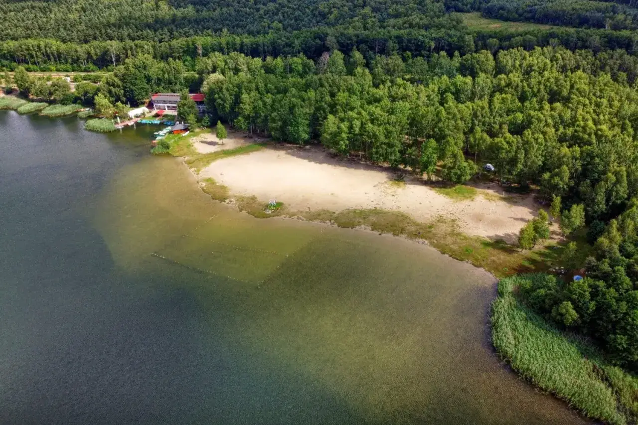 Malownicze pogoria jezioro z piaszczystą plażą, lasem i budynkami ośrodka wypoczynkowego. Woda jest przejrzysta, widać dno.