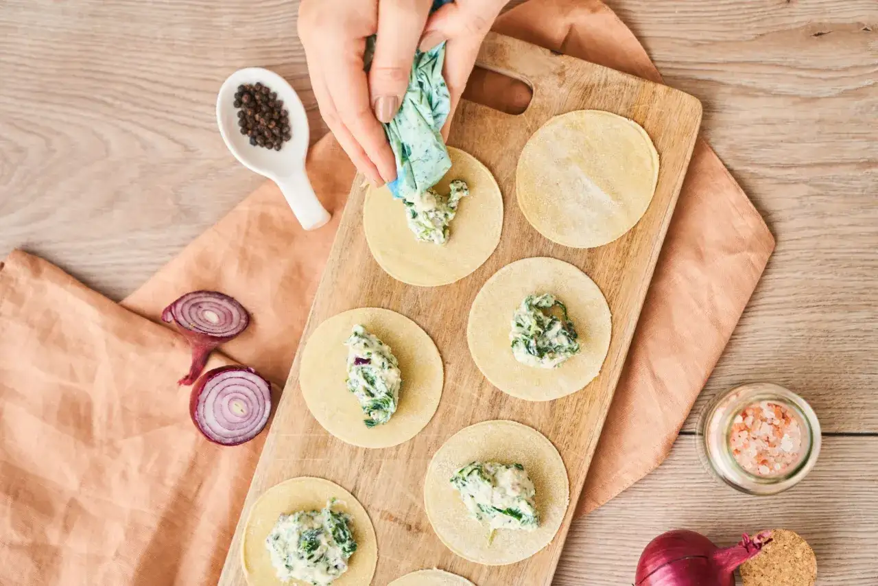 H&auml;nde f&uuml;llen Teigkreise mit einer cremigen Spinatf&uuml;llung, um Tortellini selber zu machen. Pfefferk&ouml;rner und Zwiebeln liegen bereit.