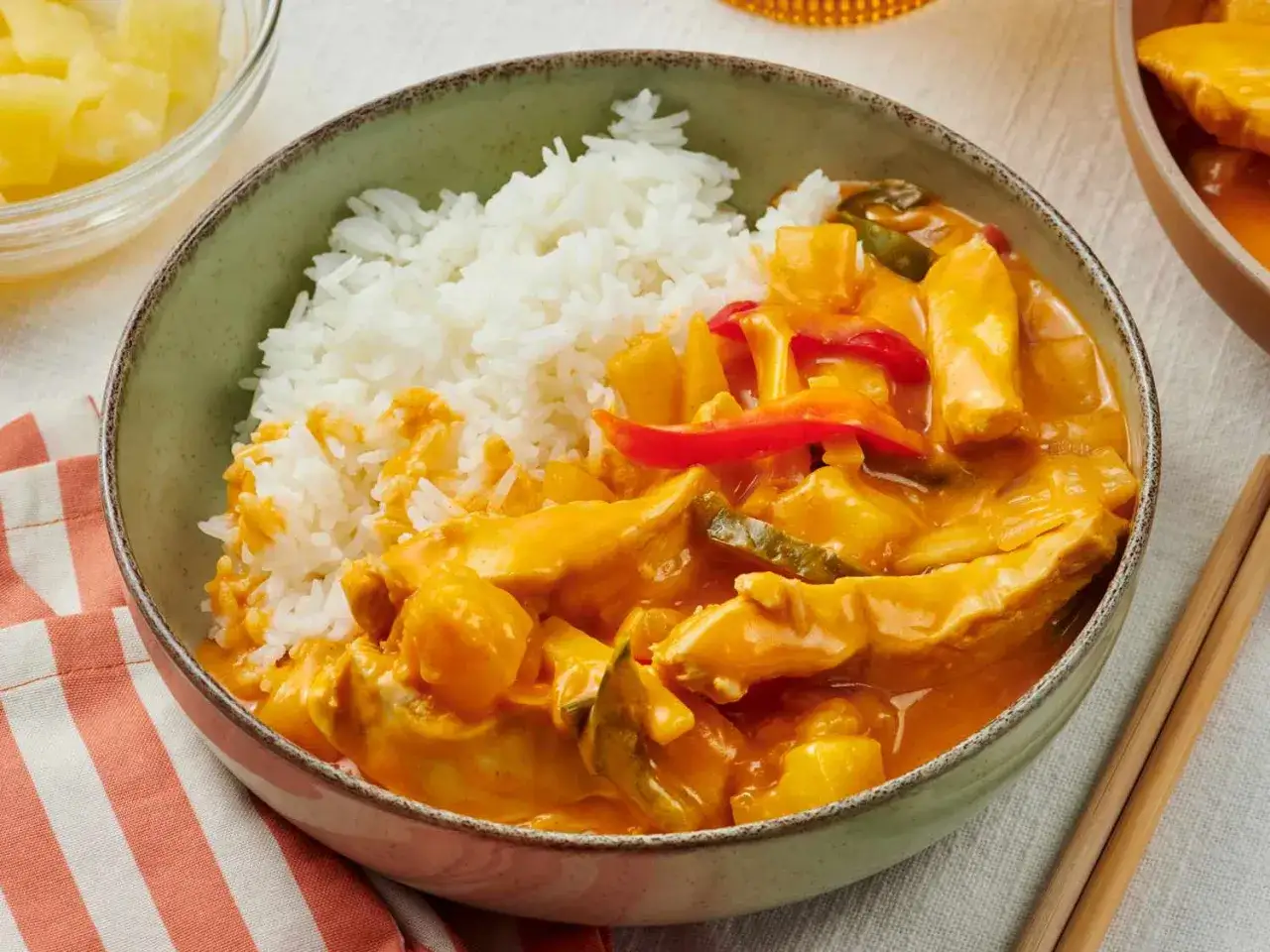 Sch&uuml;ssel mit Reis und cremigem thai curry mit ananas und kokosmilch, garniert mit Paprika und Zucchini. Daneben ein Sch&auml;lchen mit Ananasst&uuml;cken.