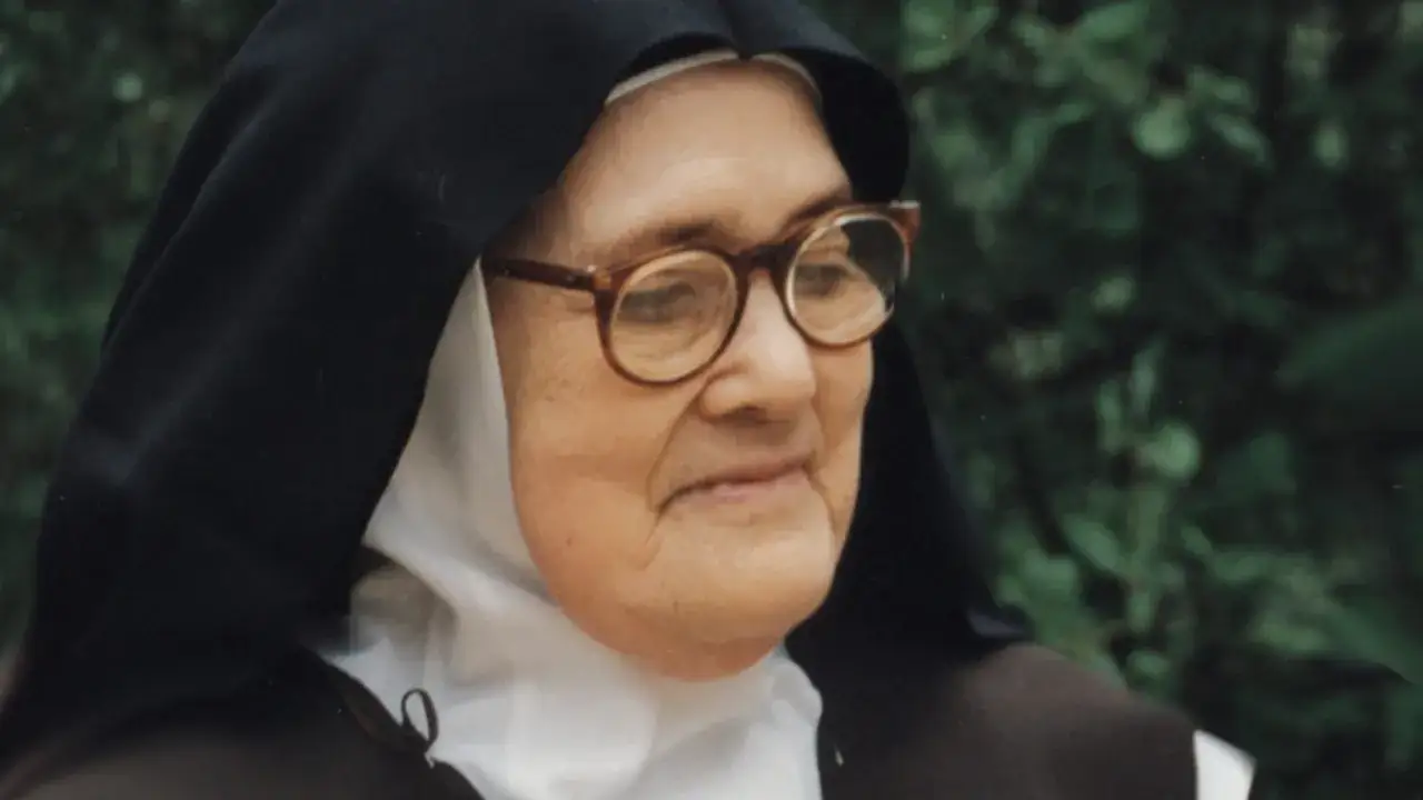 Siostra Łucja z Fatimy, w okularach i habicie, z uśmiechem na twarzy, na tle zieleni.