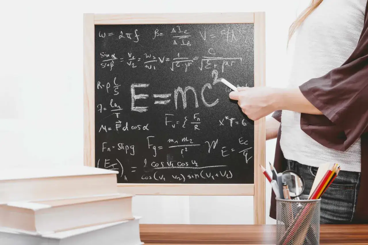 Tablica z równaniami fizycznymi, w tym słynne E=mc², które co odkrył Albert Einstein. Obok stos książek i piórnik z przyborami.