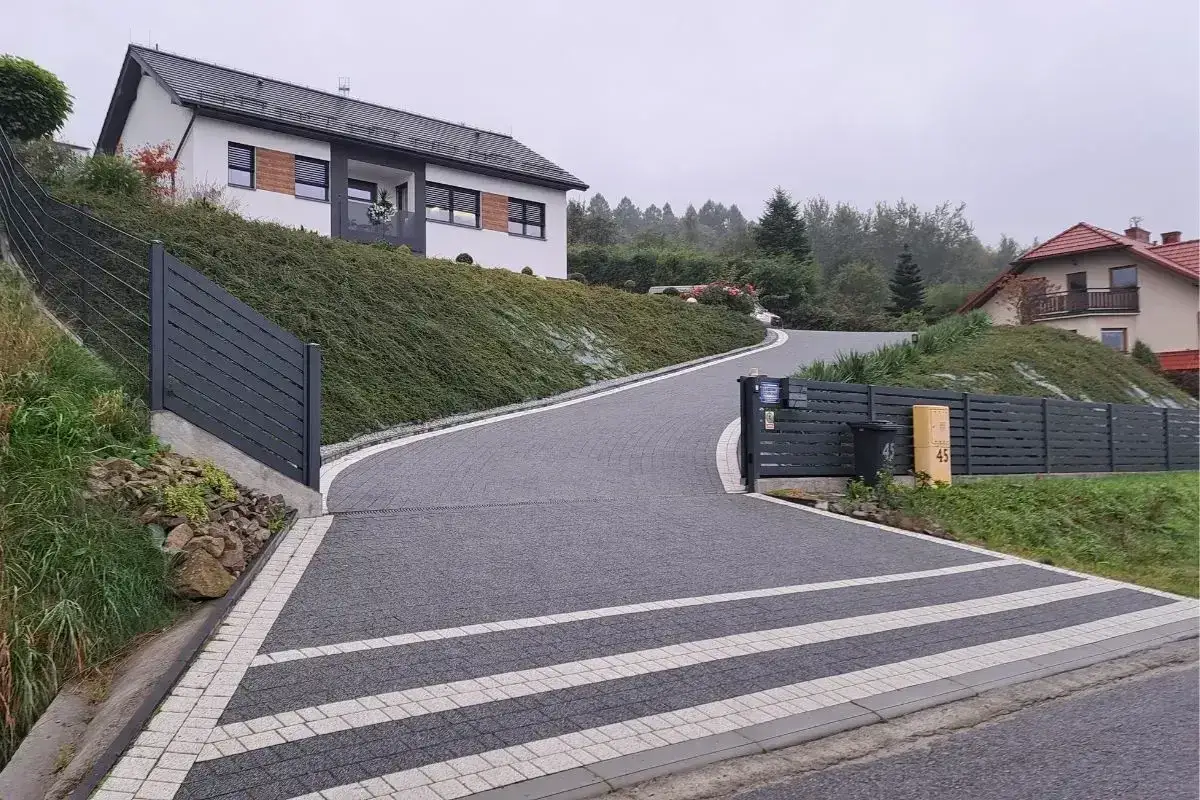 Une all&eacute;e en pente pour voiture serpente vers une maison moderne, bord&eacute;e de v&eacute;g&eacute;tation luxuriante et d'une cl&ocirc;ture sombre.
