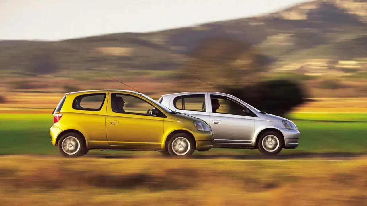 Due **toyota yaris modelli**, una gialla e una argento, sfrecciano su una strada di campagna con un paesaggio sfocato sullo sfondo.