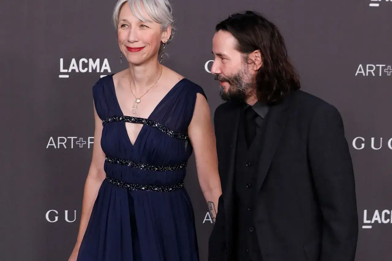 Alexandra Grant wzrost jest widoczny obok Keanu Reevesa. Oboje ubrani elegancko, ona w granatowej sukni, on w garniturze.