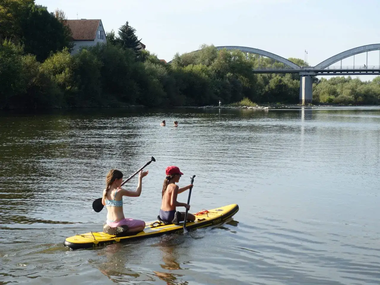 Zwei M&auml;dchen genie&szlig;en **stand up paddling** auf dem Neckar bei Heidelberg. Im Hintergrund eine Br&uuml;cke und B&auml;ume.