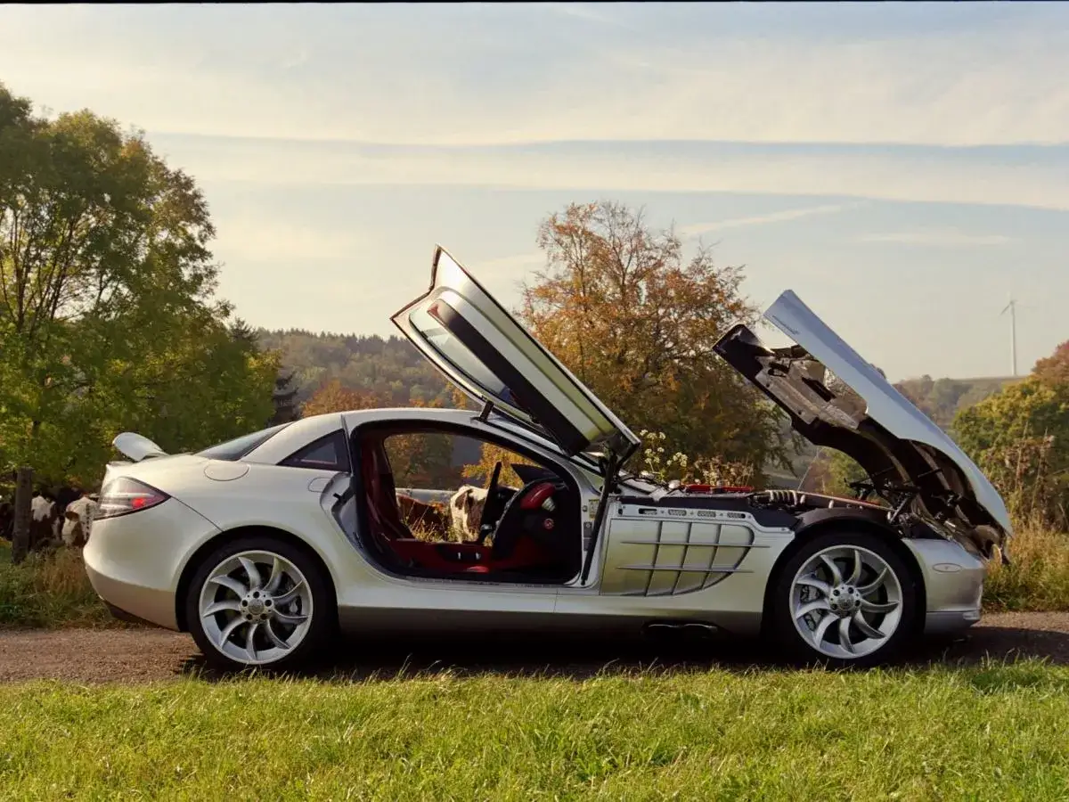 Srebrny SLR McLaren z otwartymi drzwiami i maską, zaparkowany na tle wiejskiego krajobrazu z krowami i wiatrakiem.