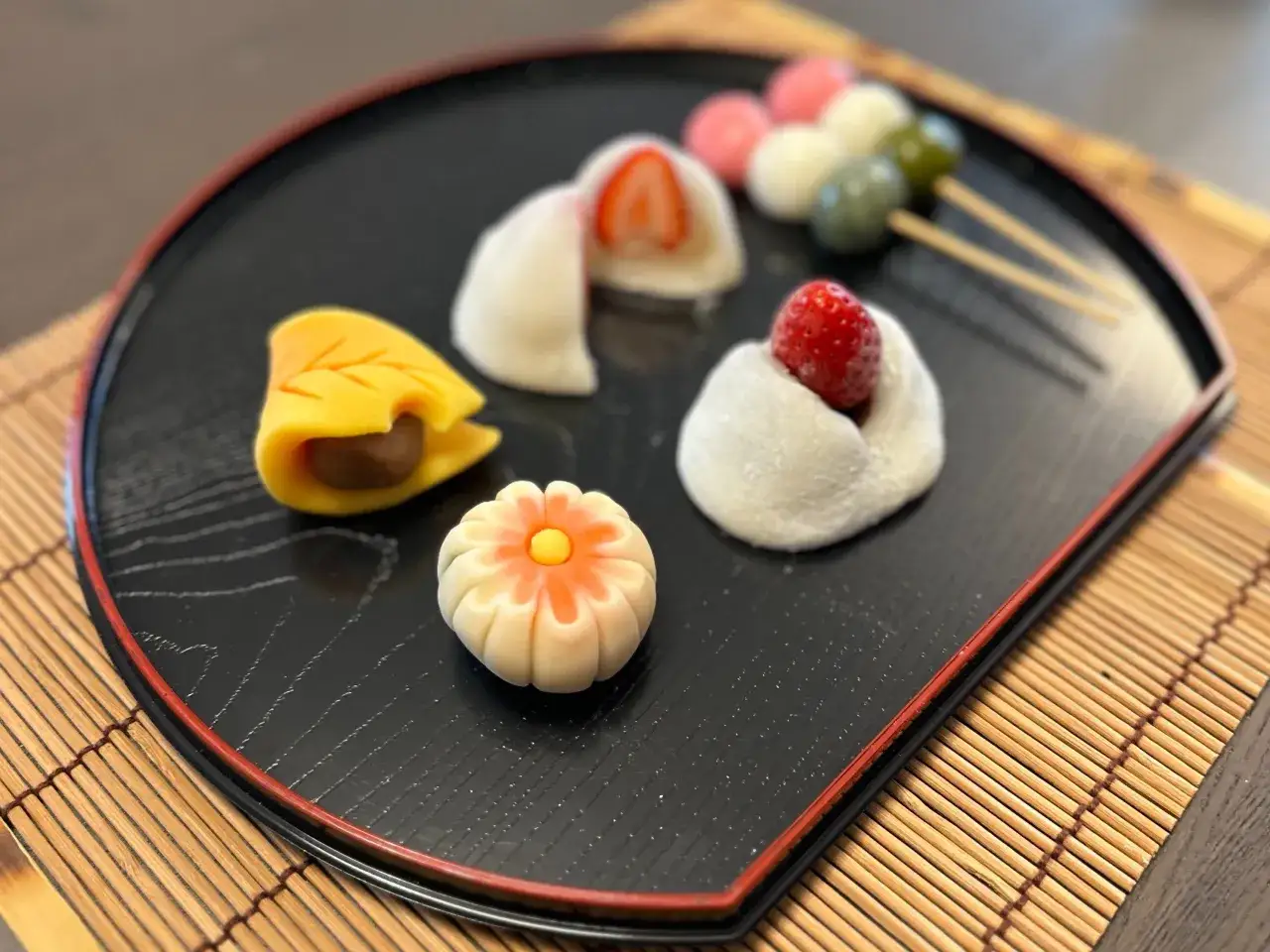 Bunte Wagashi-S&uuml;&szlig;igkeiten, inspiriert von einem Wagashi-Rezept, auf einem schwarzen Teller: Mochi mit Erdbeeren, ein Blatt mit F&uuml;llung und eine Blumenform.