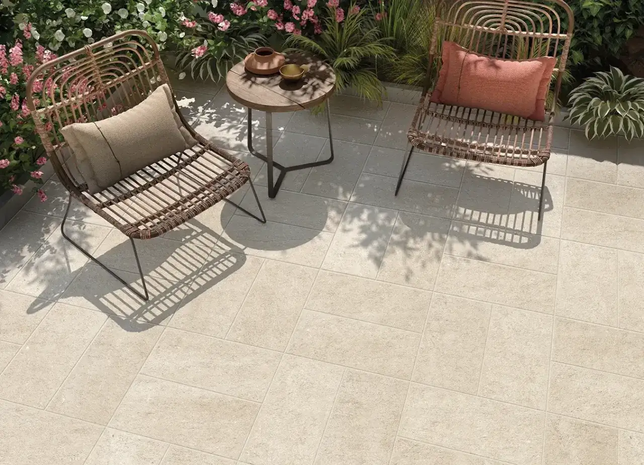 Un angolo relax su pavimenti per terrazzo in gres porcellanato. Due sedie in rattan, un tavolino e piante fiorite creano un'atmosfera accogliente.