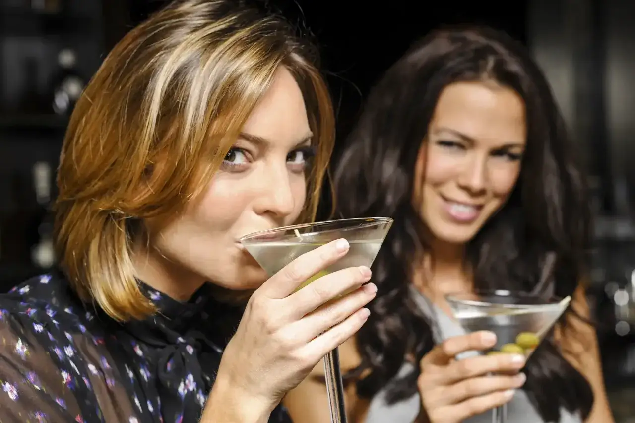 Dwie kobiety cieszą się drinkiem z wódką w barze. Jedna z nich pije martini, druga trzyma kieliszek z oliwkami.