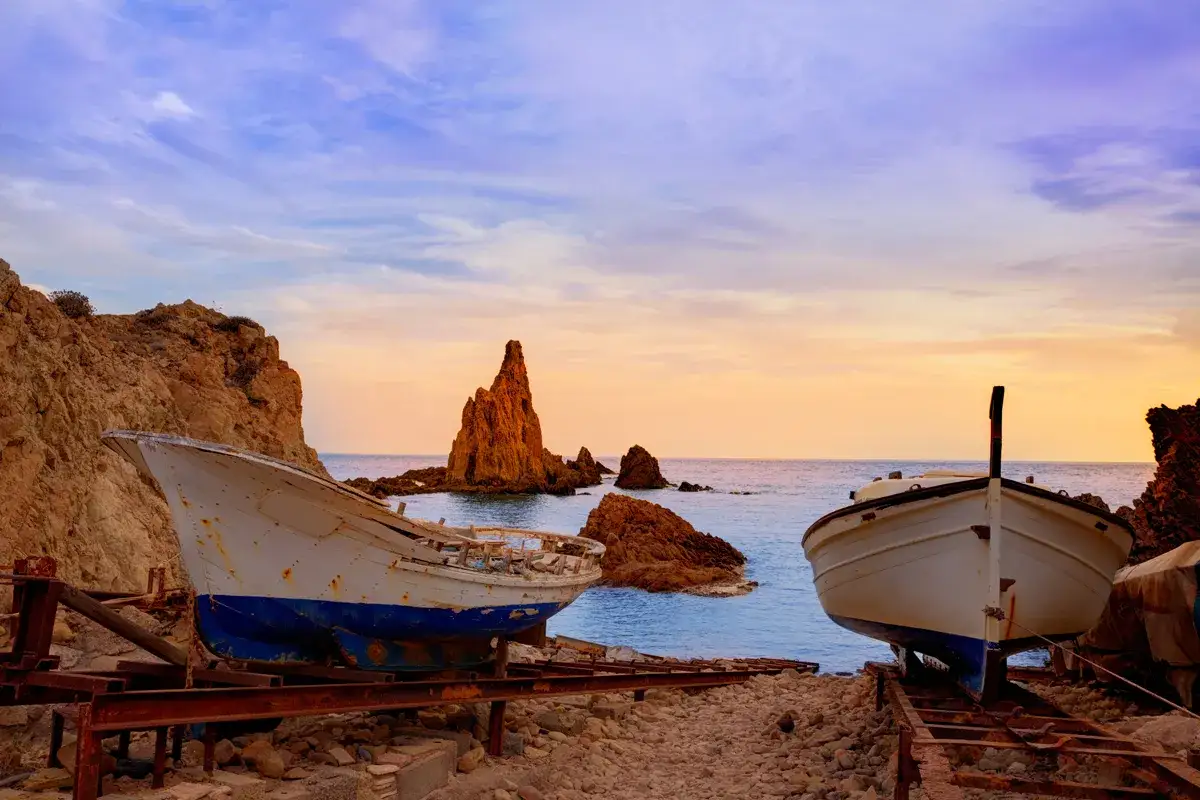 Dos barcos en la playa, con el faro de Cabo de Gata al fondo, bajo un cielo crepuscular.