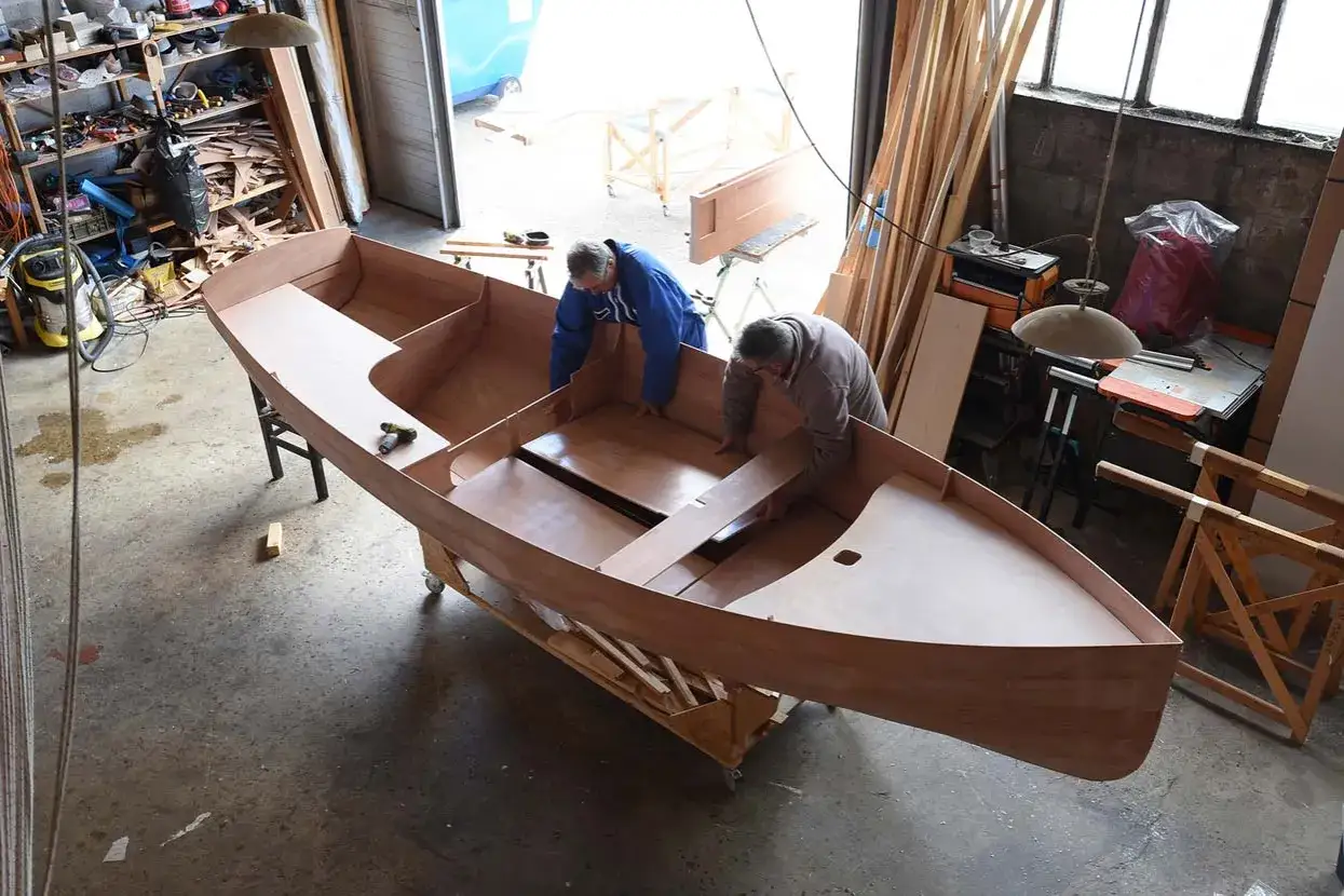 Deux hommes travaillent dans un atelier, concentrés sur la construction de leur bateau en bois. L'assemblage progresse.