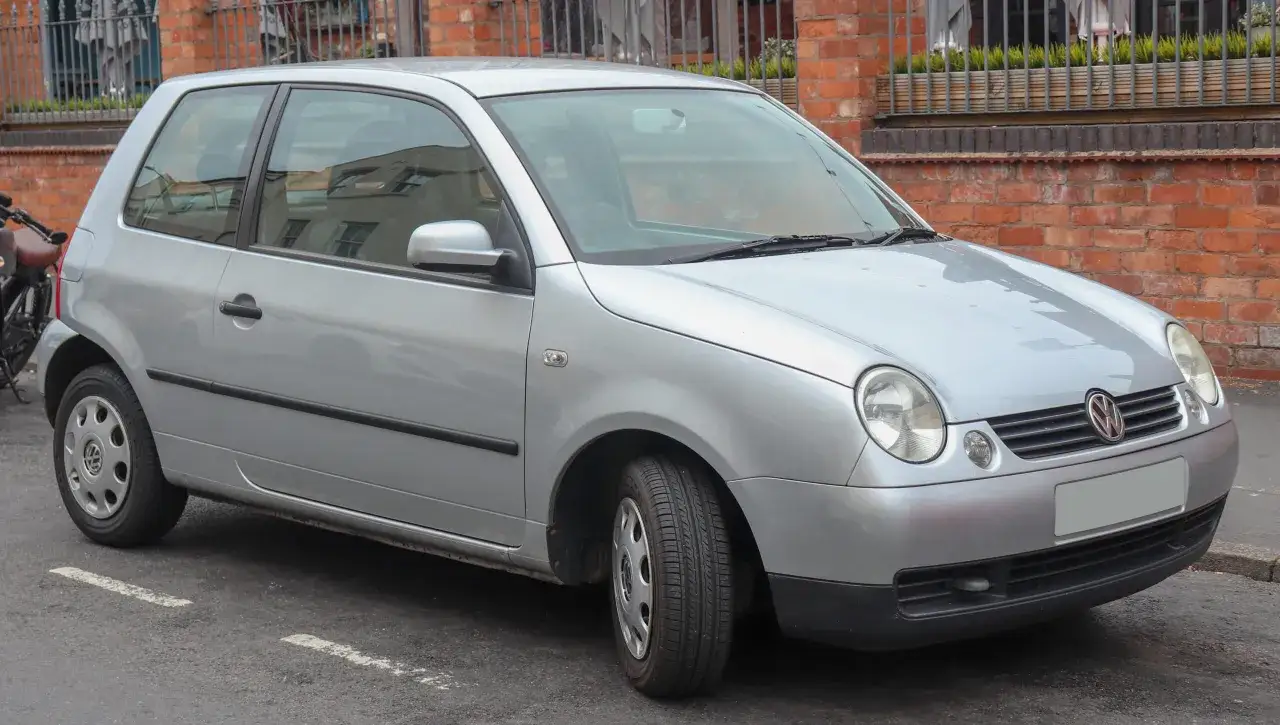 Srebrny Volkswagen Lupo stoi zaparkowany przy ceglanej ścianie.