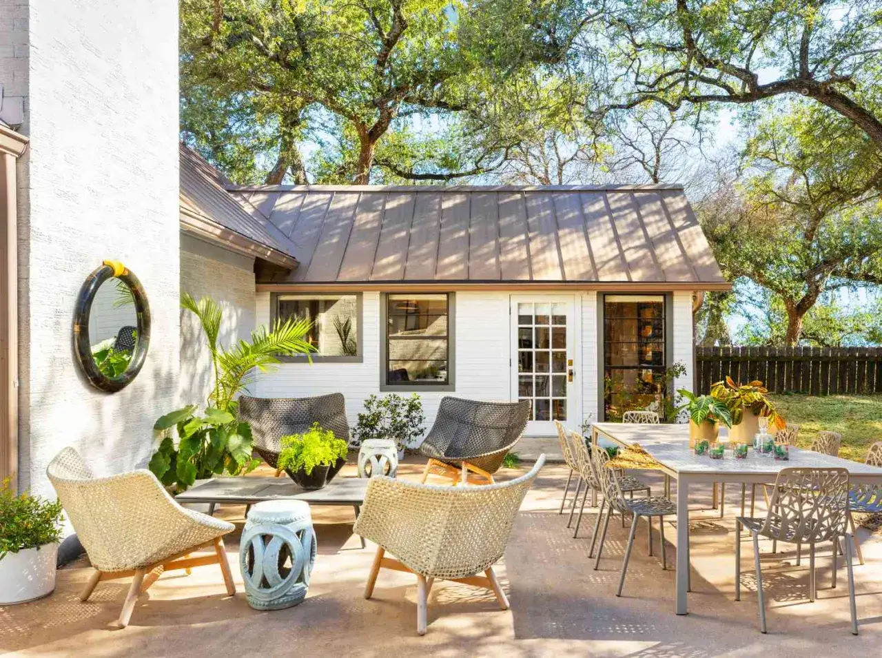Acogedor patio con muebles de mimbre y metal, plantas exuberantes y una encantadora cottage casa al fondo, bajo la sombra de &aacute;rboles frondosos.