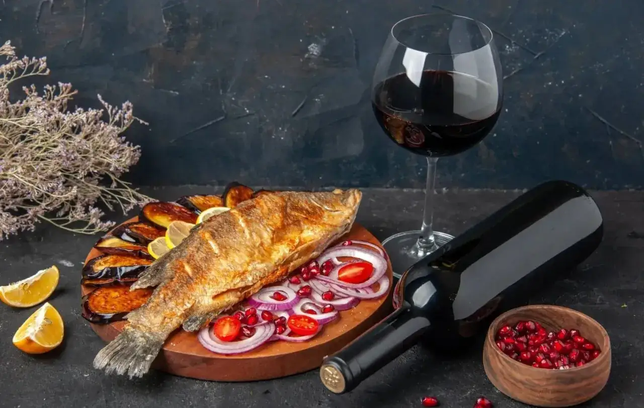 Piatto di pesce fritto con melanzane, limone e cipolla rossa, accompagnato da un bicchiere e una bottiglia di vino rosso da abbinare al pesce.