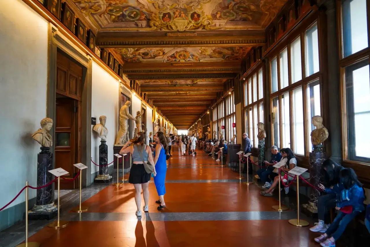 Wnętrze muzeum Uffizi z rzeźbami i malowanym sufitem. Ludzie spacerują i odpoczywają w galerii.