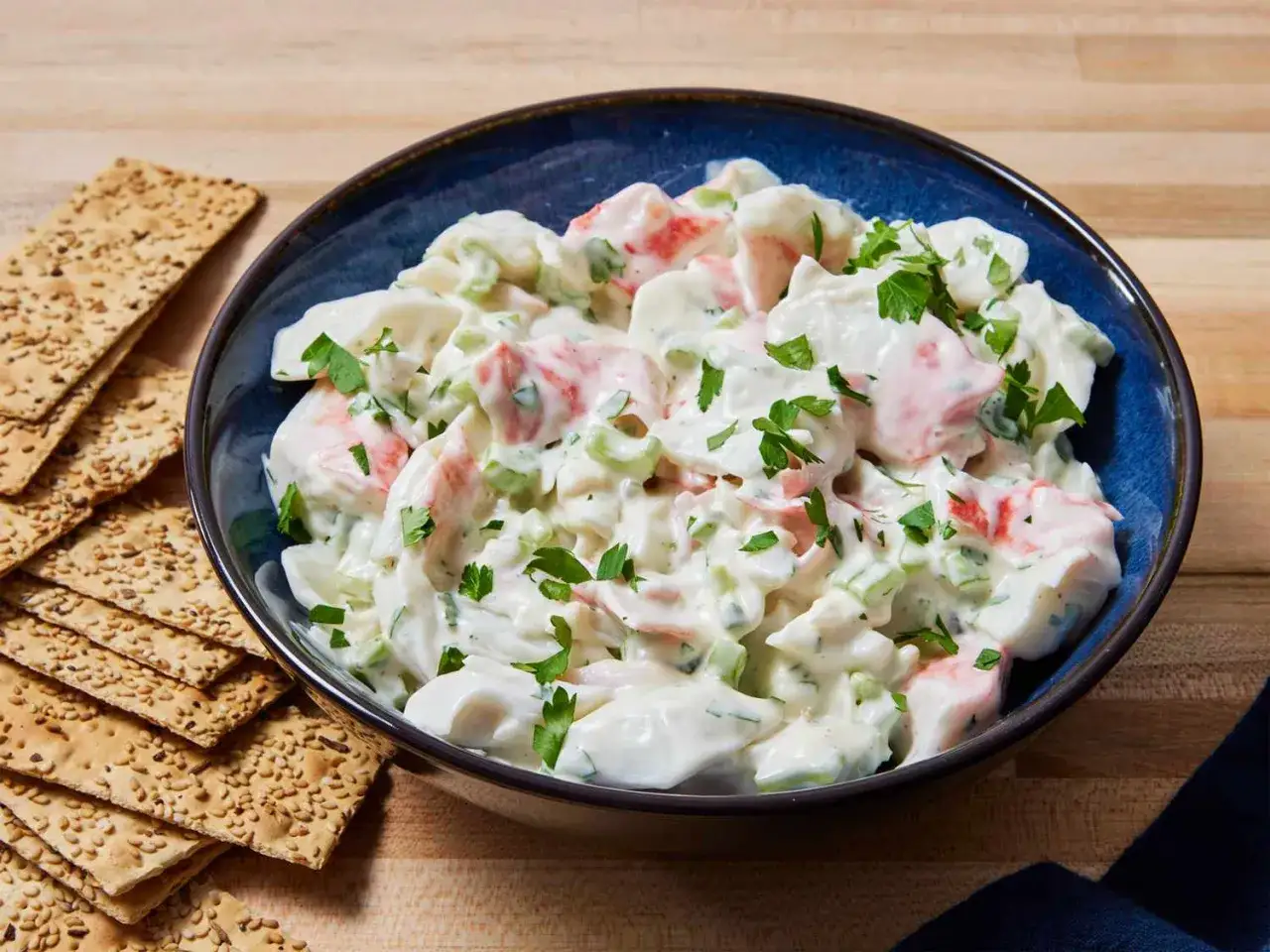 Deliciosa ensalada de cangrejo cremosa, adornada con perejil fresco, servida en un taz&oacute;n azul junto a galletas crujientes.