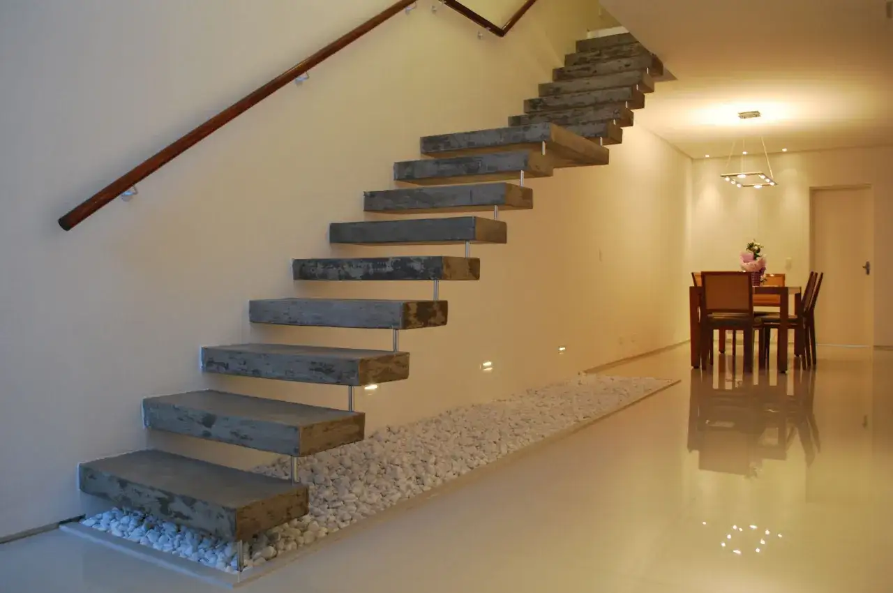 Escaleras r&uacute;sticas modernas con pelda&ntilde;os de hormig&oacute;n flotantes y barandilla de madera. Un camino de guijarros blancos bordea la base.