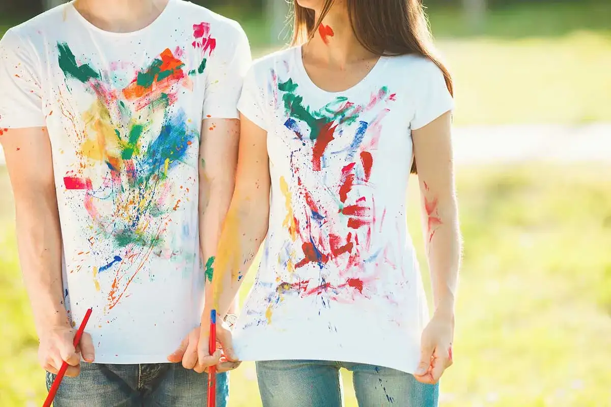 Bunte Farbspritzer auf wei&szlig;en T-Shirts. Ein Paar h&auml;lt Pinsel, bereit, die Textilfarbe zu entfernen und neue Kunst zu schaffen.