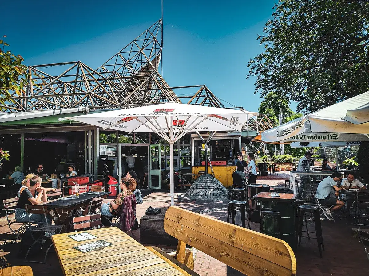 Sonniger Biergarten im Schlossgarten mit G&auml;sten an Tischen unter Sonnenschirmen.