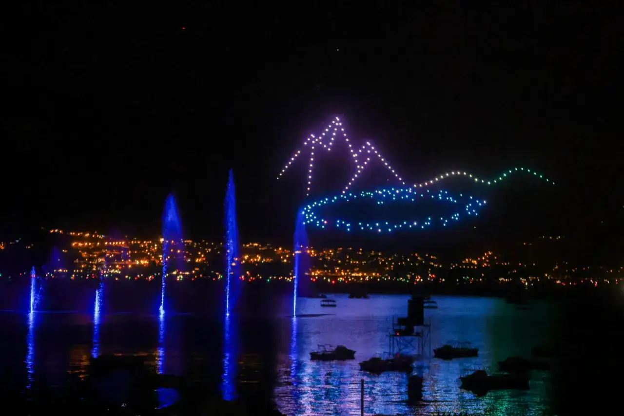 Spectacle lumineux de drones formant des montagnes et des vagues au-dessus d'un lac, avec des fontaines bleues illuminant l'eau. Une belle fete du lac.