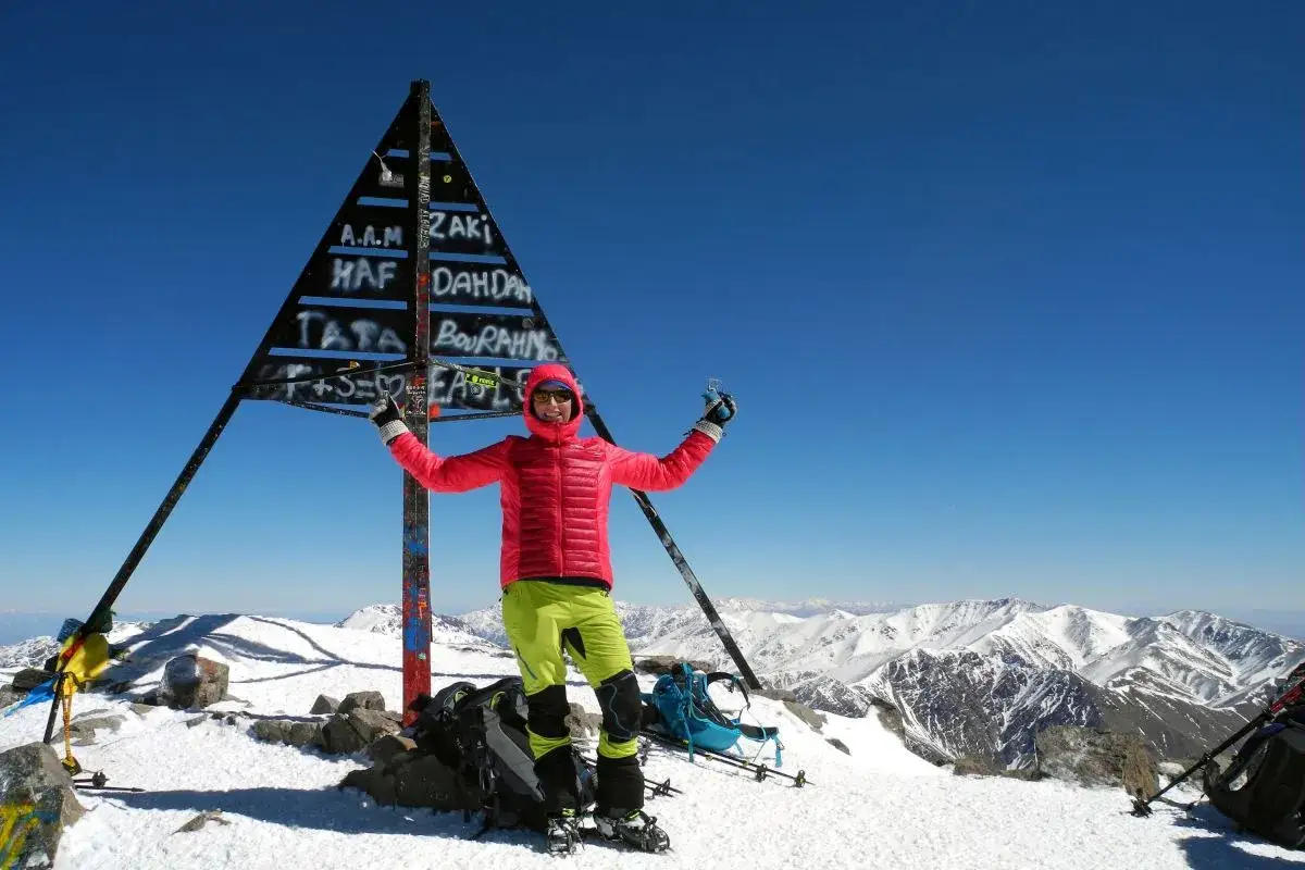 Erfolgreiche Gipfelbesteigung am Jbel Toubkal! Wanderer jubelt vor dem Gipfelzeichen in schneebedeckter Berglandschaft.