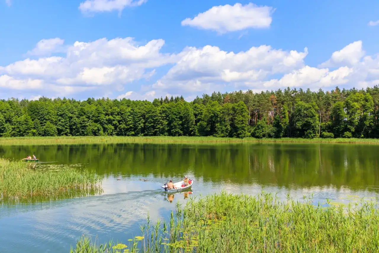 Spokojny dzień na Mazurach. Ludzie pływają łódką po jeziorze, otoczeni zielenią lasów. Idealne miejsce na wypoczynek, jak w urokliwych miasteczkach na Mazurach.
