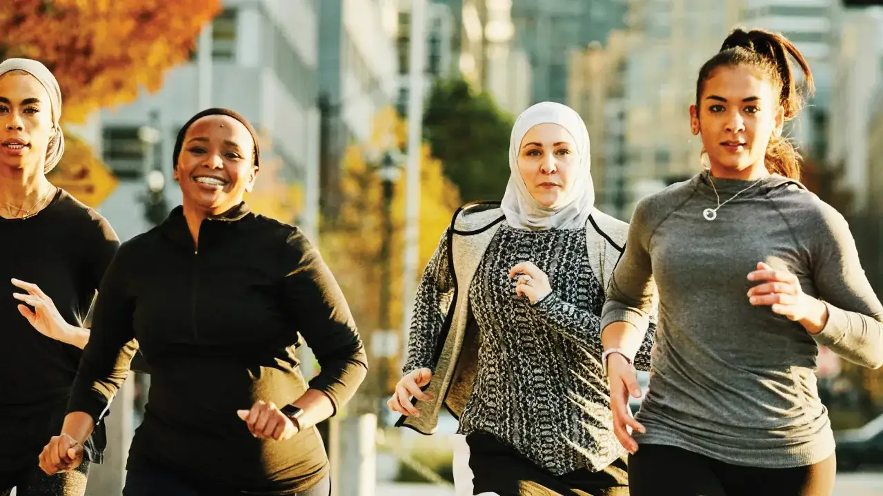 Vier Frauen joggen in der Stadt. Sie lachen und genießen die Bewegung. Das ist, was bringt joggen: Freude und Gemeinschaft.