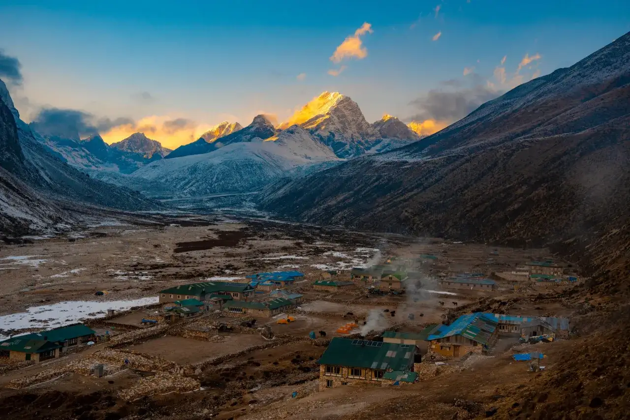 Wioska u podn&oacute;ża ośnieżonych szczyt&oacute;w. Dym unosi się z dom&oacute;w, sugerując ciepło i życie. Planujesz wyprawę? Sprawdź **Everest Base Camp koszty**.