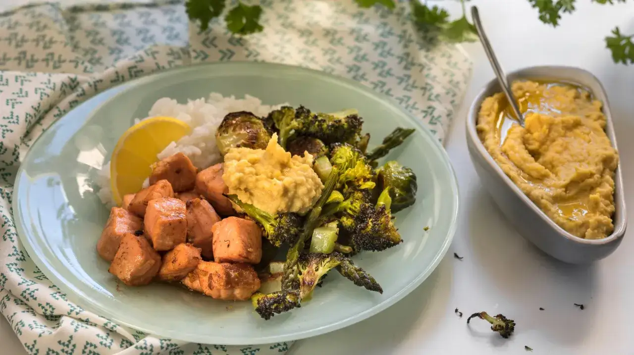 En tallrik med nyttig mat f&ouml;r att g&aring; ner i vikt: laxbitar, ris, ugnsrostad broccoli och brysselk&aring;l, samt en sk&aring;l hummus.