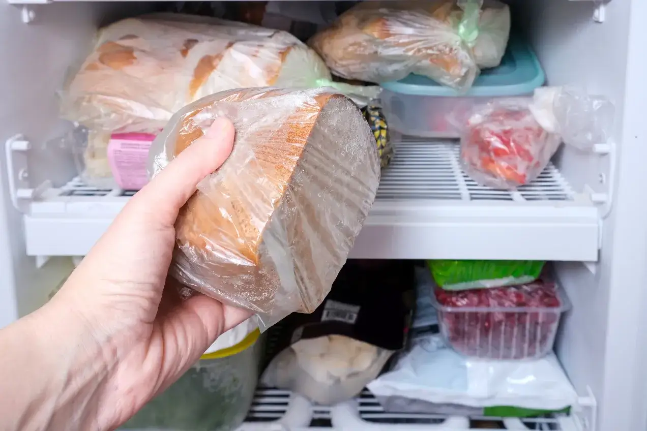 Ręka wyjmuje z zamrażarki bochenek chleba w folii. W zamrażarce widać inne produkty. Ile można trzymać chleb w zamrażarce? Nawet kilka miesięcy!