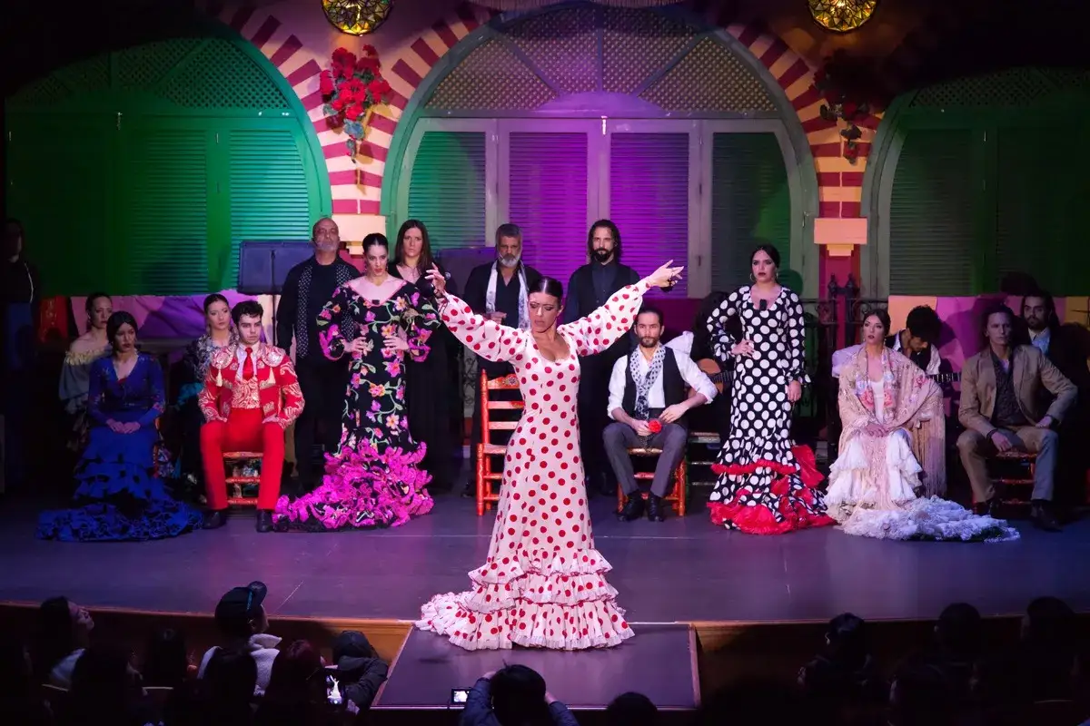 Un grupo de artistas de flamenco, con trajes vibrantes, incluyendo un vestido de lunares rojos y blancos, bailan en un escenario.