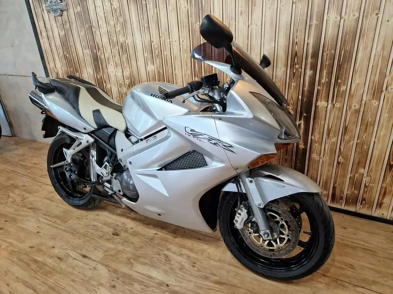 Srebrny motocykl Honda VFR 800 z dwukolorowym siedzeniem, gotowy do jazdy po drewnianej podłodze.