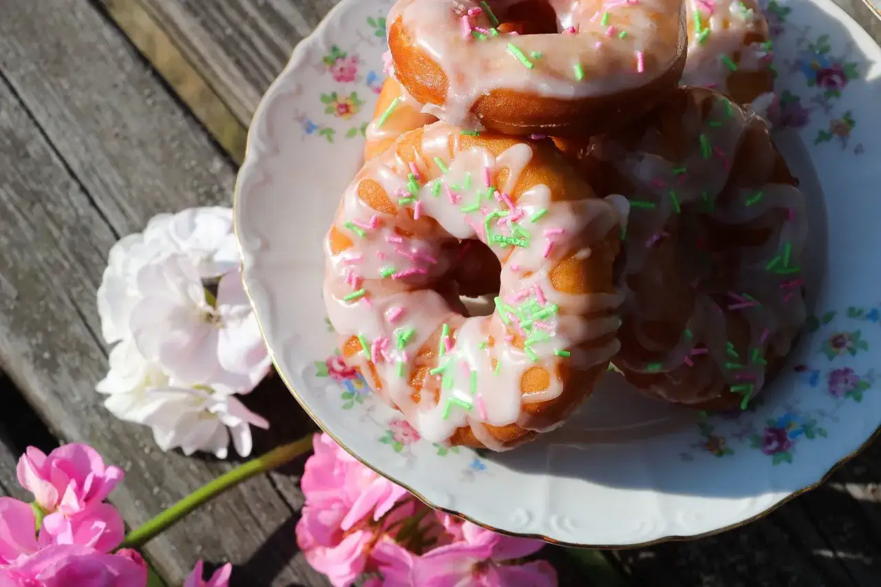 En hög med hembakade donuts med rosa och grön strössel på en blommig tallrik. Perfekt för en fika utomhus.