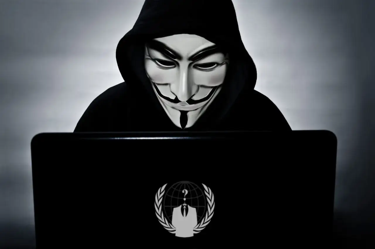 Osoba w masce Guya Fawkesa, symbol grupy anonymous, pochyla się nad laptopem z charakterystycznym logo.