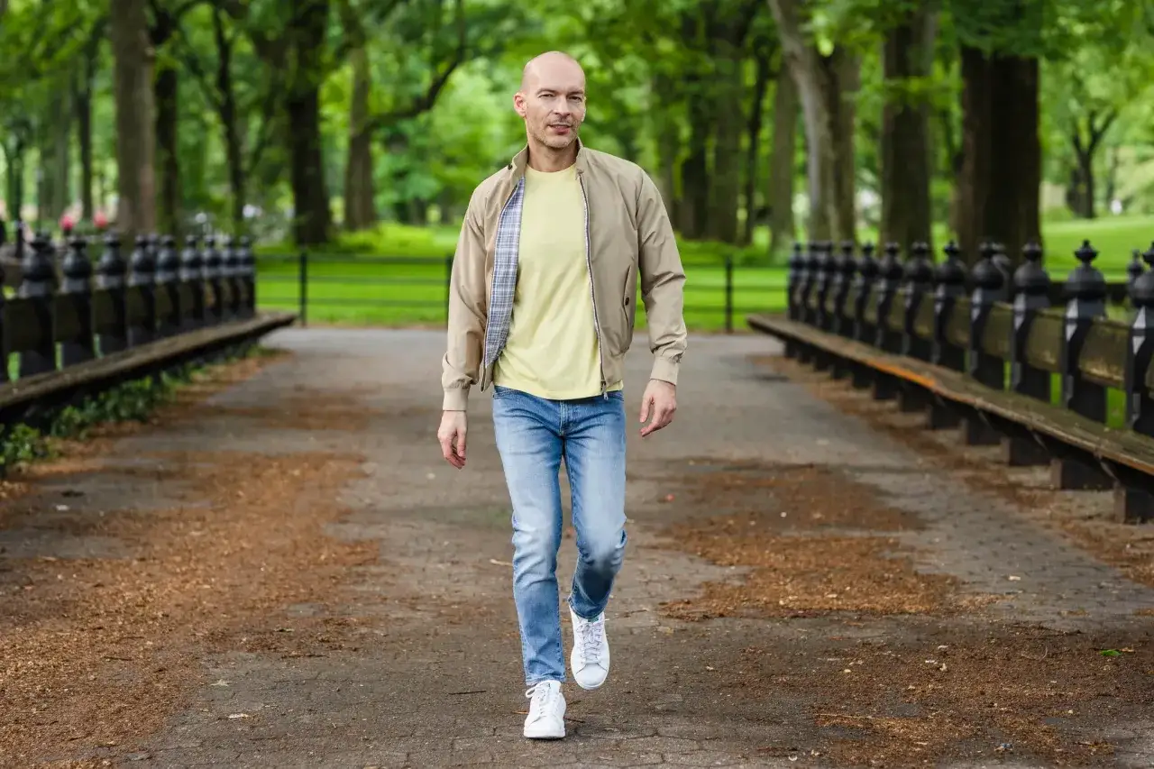 Mann im Park, der ein l&auml;ssiges Outfit zusammenstellt: beige Bomberjacke, gelbes T-Shirt, Jeans und wei&szlig;e Sneaker.