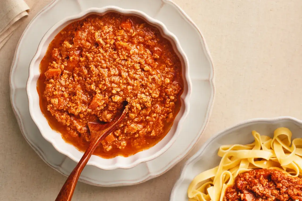 Gęsty sos mięsny, idealny do spaghetti bolognese przepis oryginalny, w miseczce z drewnianą łyżką. Obok porcja tagliatelle.
