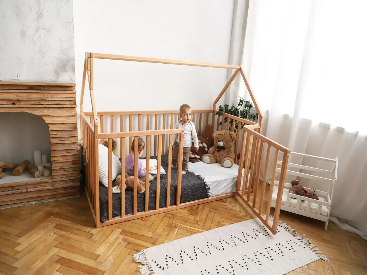 Two children play in a wooden playpen floor bed, one reading a book, the other standing. A teddy bear sits nearby.