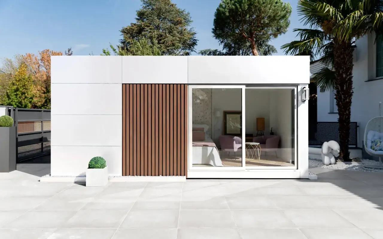 Moderna casa con techos bajos, paredes blancas y detalles de madera. Se ve el interior con una cama y sillones.