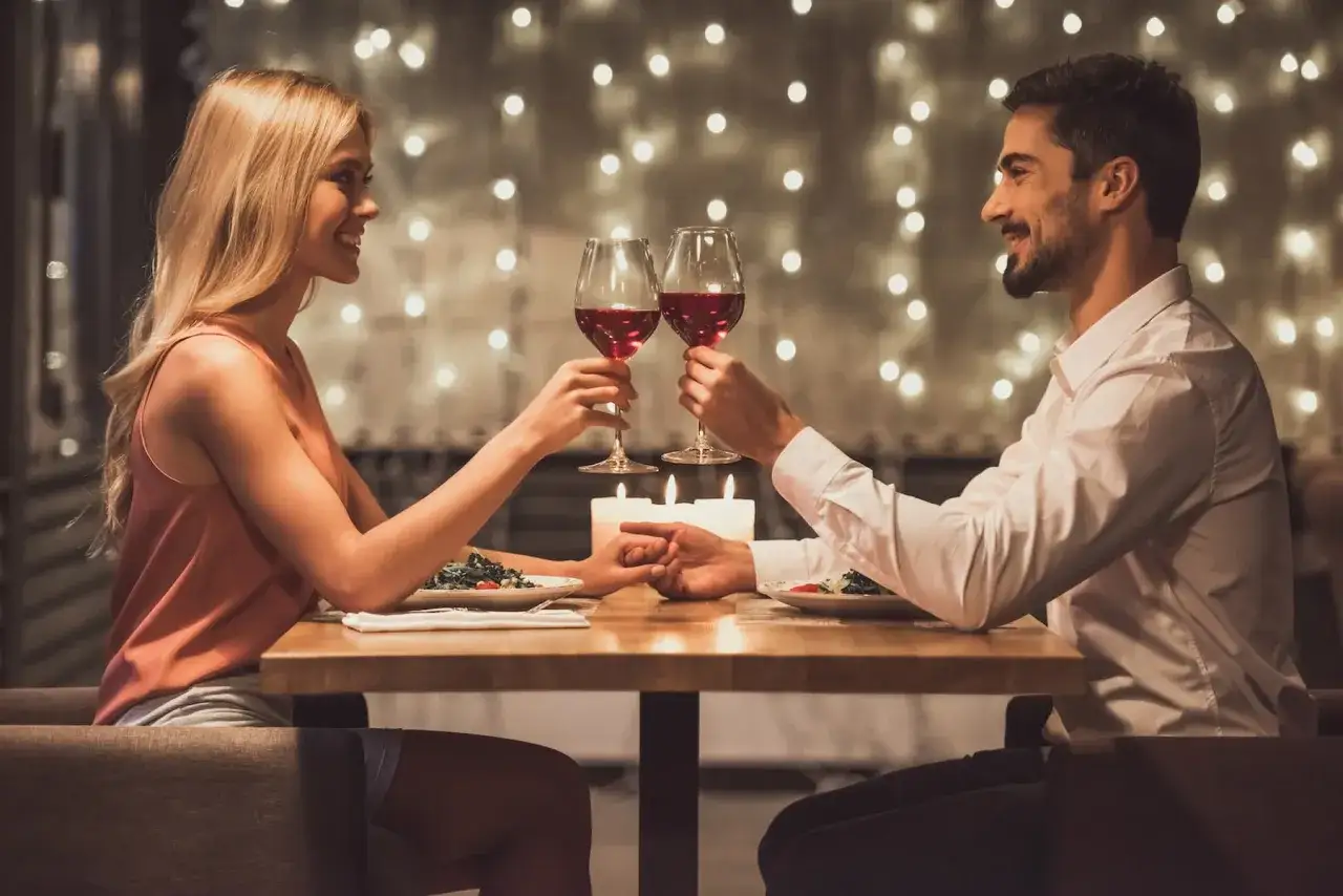 Para młoda wznosi toast kieliszkami wina podczas romantycznej kolacji. Ich dłonie splecione pod stołem, a w tle migoczą światełka. Idealne pierwsze randki.