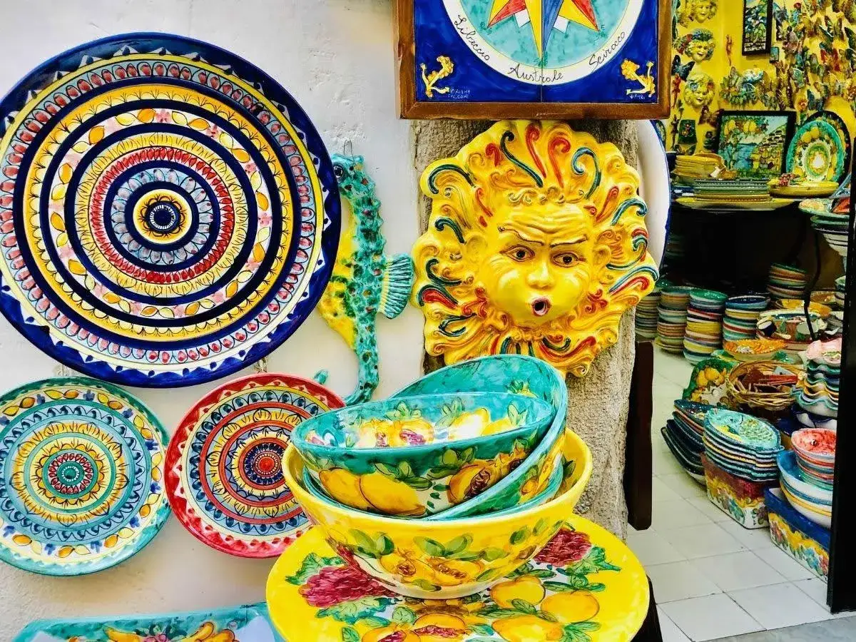 Vibrant ceramic art, including a sun face and seahorse, adorns a shop in Vietri sul Mare. Colorful plates and bowls overflow with fruit designs.