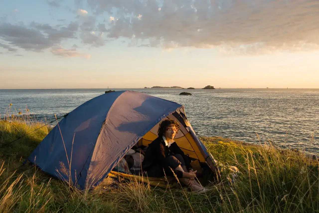 Un bivouac GR34 au coucher du soleil. Une personne est assise dans une tente bleue face à la mer.