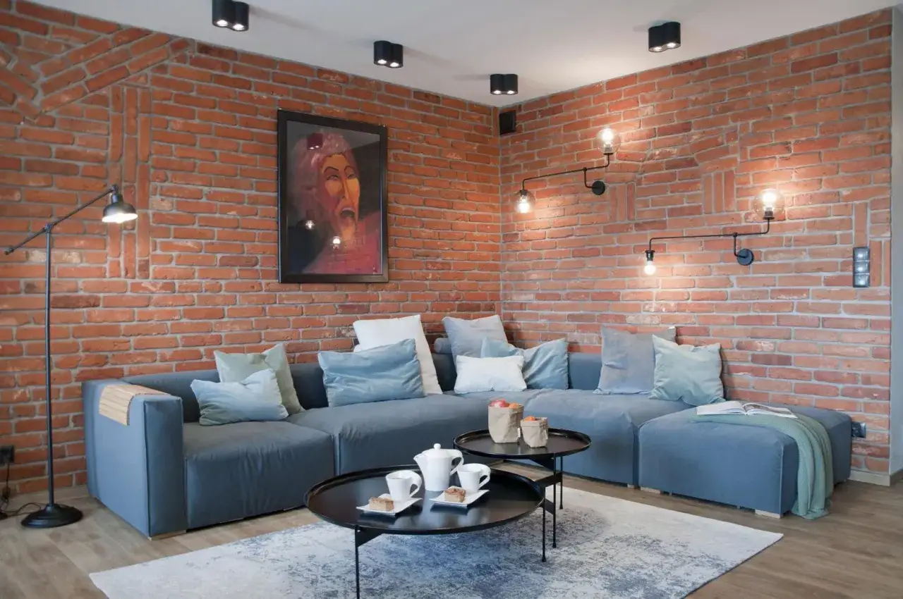 Nowoczesny salon z ceglaną ścianą i szarą sofą. Wygodne poduszki i stoliki kawowe tworzą przytulną atmosferę, a punkty świetlne dodają charakteru.