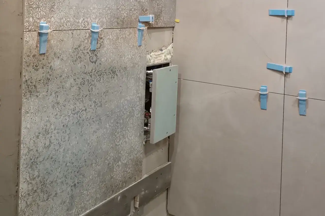Montaż płytek łazienkowych z widocznymi klinami. W ścianie otwór z instalacją, przykryty częściowo drzwiczkami rewizyjnymi 60x60.