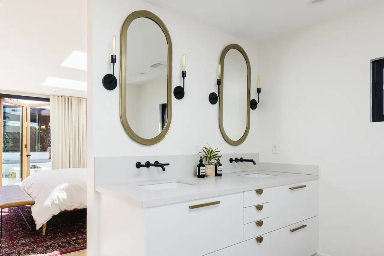 Modern double vanity bathroom ideas: two oval mirrors with gold frames, black sconces, white countertop, and white cabinets with brass hardware.