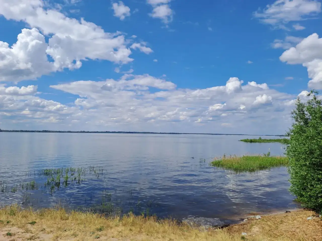 Spokojny zalew Jeziorsko w letni dzień. Błękitne niebo z białymi chmurami odbija się w tafli wody.