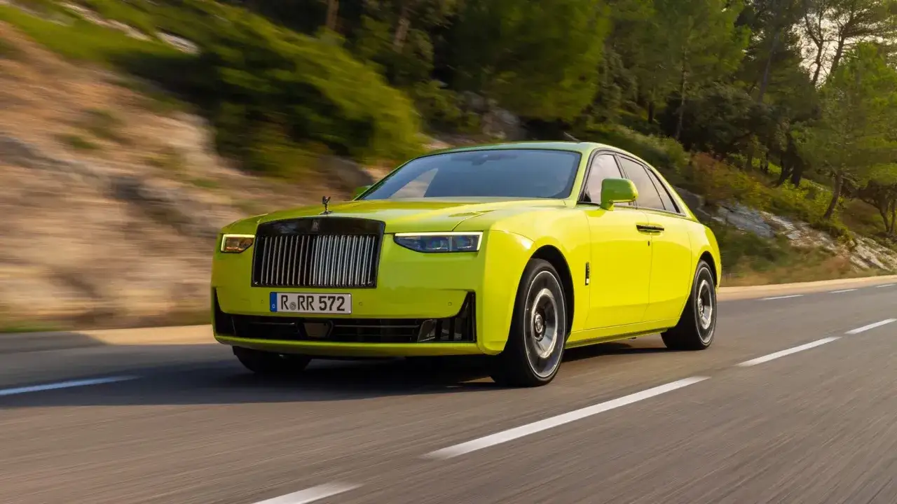 Jaskrawożółty Rolls-Royce Ghost sunie po krętej drodze wśród zielonych wzgórz.