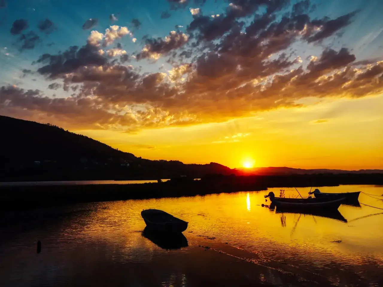 Atardecer dorado sobre aguas tranquilas en Cantabria. Barcas reposan en la orilla, reflejando el cielo anaranjado. Un paisaje sereno de playas limpias Cantabria.